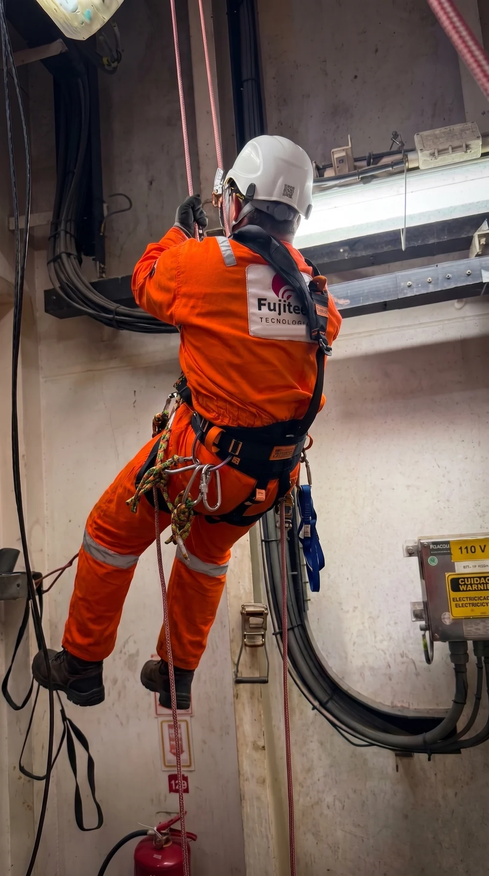 Técnico da Fujitec com macacão laranja trabalhando em altura com acesso por corda, junto a eletrocalhas e uma caixa elétrica